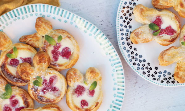 Plates of bunny-shaped pastries filled with custard and raspberries, garnished with mint leaves, rest atop a light wooden surface, conveying a festive and inviting atmosphere.