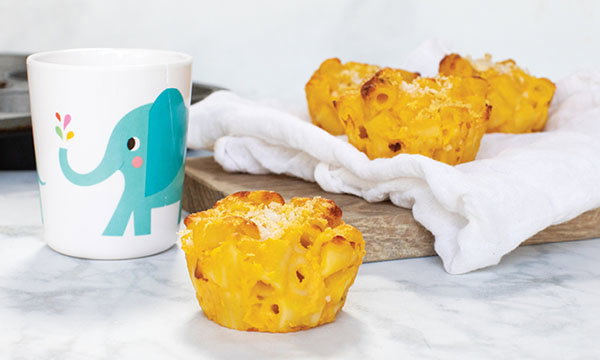 A cute elephant mug sits beside golden mac and cheese muffins on a wooden board with a white cloth, creating a cosy and playful atmosphere.
