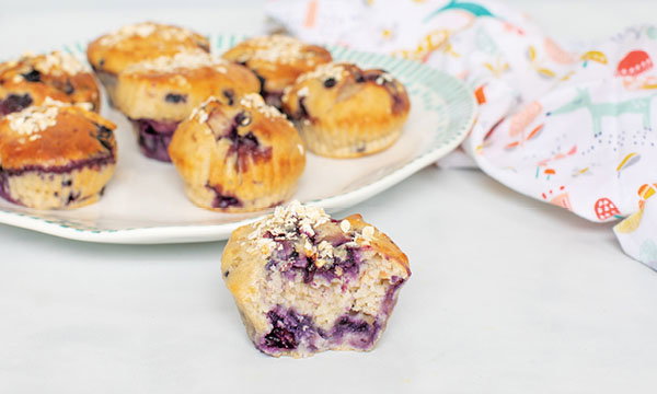 Plate of blueberry muffins topped with oats, set on a white table. One muffin with a bite taken out sits in front, with a colorful cloth nearby.