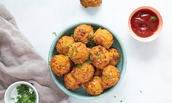 A bowl of golden, crispy sweet potato and carrot bites garnished with herbs, accompanied by a small dish of red sauce, set on a light surface with a cloth napkin.