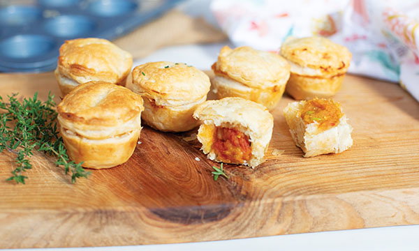 Mini savory samosa pies with golden flaky crusts rest on a wooden board. One is bitten, showing orange filling. Fresh thyme and a napkin are nearby.