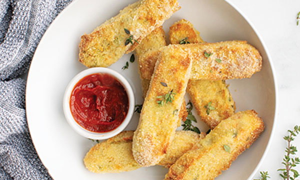 Golden mixed vegetable fingers on a white plate, sprinkled with herbs, served with a small bowl of red marinara sauce. A textured grey napkin is nearby.