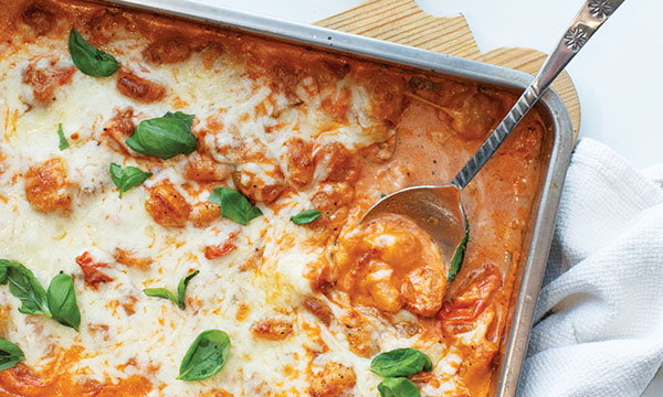 Baked gnocchi with melted cheese and marinara sauce in a metal tray, garnished with fresh basil leaves. A spoon rests in the dish, conveying warmth.