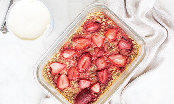 A glass dish filled with strawberry oatmeal bake, topped with sliced strawberries, sits on a marble surface. A bowl of yogurt and a white cloth are nearby.