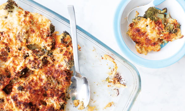 A glass baking dish with cheesy broccoli casserole, partly served. A portion sits in a blue-rimmed bowl. The dish looks golden and crispy.