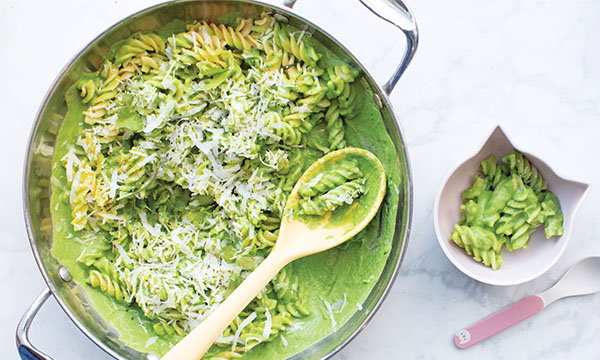 A skillet of creamy green pasta topped with shredded cheese, next to a small bowl of the same pasta. A wooden spoon rests in the skillet, conveying a fresh, appetising mood.
