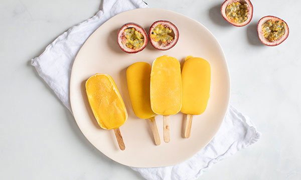 Four yellow popsicles on a white plate with two halved passion fruits, set on a white napkin. Bright and refreshing summer vibe.