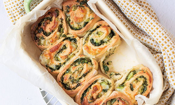 Golden-brown spinach and cheese rolls in a parchment-lined tray on a white surface. The rolls are arranged neatly, exuding a warm, appetising look.