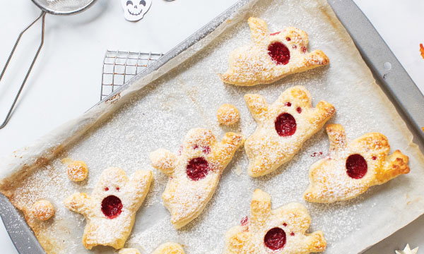 Ghost-shaped jam pastries dusted with icing sugar and filled with red jam, arranged on a baking tray with small pastry cut-outs
