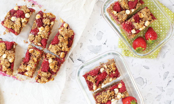 Sliced raspberry oat bars with white chocolate chips sit on parchment paper and in a glass dish. Fresh strawberries add a colourful, fresh touch.