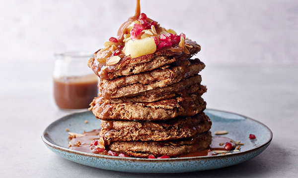 Stack of vegan pancakes topped with syrup, diced apples, pomegranate seeds, and sliced almonds on a blue plate. A small jar of sauce is blurred in the background.