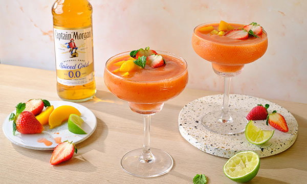Fruity strawberry mango mocktails in elegant glasses on a light surface, garnished with fresh fruit slices and mint. A spiced 0.0% rum bottle in the background.