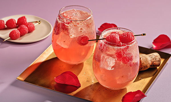 Two pink cocktails with raspberries and ice cubes in glasses on a gold tray. Rose petals and skewered raspberries add elegance on a pink surface.