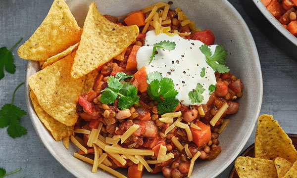 A colourful dish of chilli with beans, lentils, and carrots topped with sour cream, cheddar, and coriander, served with tortilla chips.