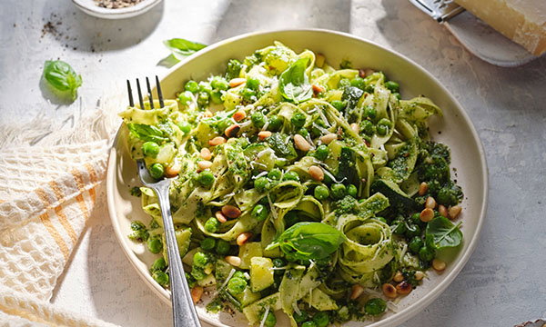 A plate of tagliatelle with vibrant pesto sauce, peas, and pine nuts, garnished with fresh basil. The scene is bright, evoking a sense of freshness.