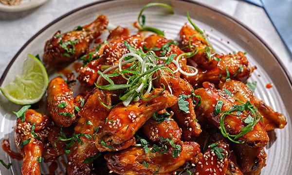 A plate of glossy, spicy chicken wings garnished with sesame seeds and fresh herbs. Lime wedge on plate adds brightness. A savoury, vibrant dish.