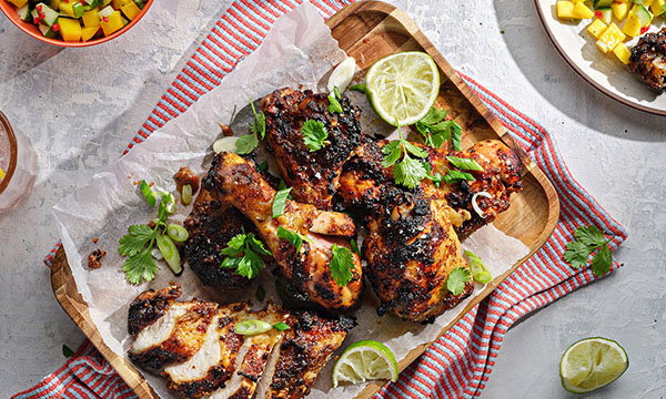 Grilled chicken with crispy skin on a wooden tray, garnished with lime slices and coriander. A vibrant, summer meal on a red-striped cloth.
