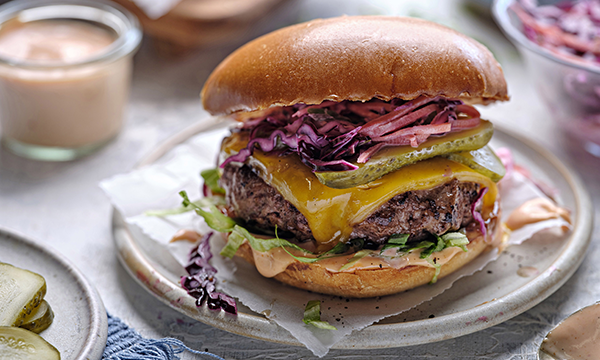Juicy cheeseburger with cheddar, pickles, shredded purple cabbage, and lettuce on a smooth bun, served on parchment-lined plate, evoking a fresh, savory vibe.