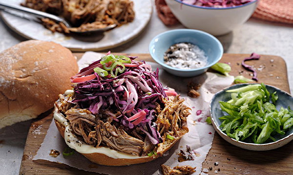 Pulled pork sandwich topped with vibrant purple coleslaw and green onions on a wooden board, accompanied by dishes of green onions and spices.
