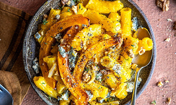 A rustic bowl of pasta topped with roasted squash, walnuts, and blue cheese crumbles, garnished with thyme. The dish is vibrant and autumnal.