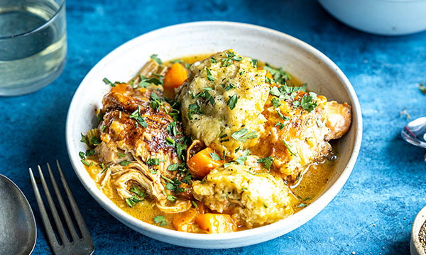 A comforting bowl of chicken and dumplings, featuring tender chicken, carrots, and fluffy dumplings in rich broth. Garnished with fresh herbs, set on a blue background.