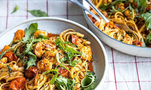 A bowl of spaghetti with shrimp, cherry tomatoes, and fresh rocket, next to a pan of the same dish on a checked cloth, conveying a fresh, hearty meal.