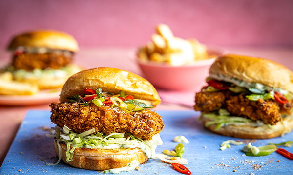 Close-up of two crispy chicken burgers with lettuce and red peppers on a blue board. A bowl of fries is blurred in the pink-toned background.