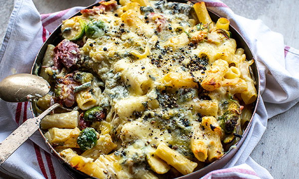 Baked pasta with melted cheese, crispy top, and speckles of black pepper in a pan, surrounded by a white cloth with red stripes and a spoon. Cosy and inviting.