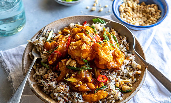 A bowl of rice topped with golden, saucy chicken, garnished with red peppers, green onions, and chopped peanuts, set on a neutral cloth with a fork and spoon.