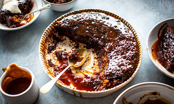 A half-eaten sticky toffee pudding in a dish, surrounded by bowls with portions served, a spoon, and rich toffee sauce, creating a cosy, indulgent scene.