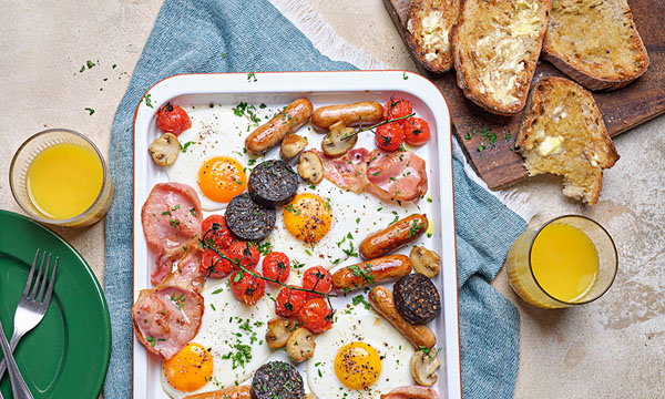 A tray of a full Irish breakfast includes fried eggs, sausages, bacon, tomatoes, mushrooms, and black pudding. Slices of buttered toast and two glasses of orange juice are set nearby, creating a hearty and inviting meal.