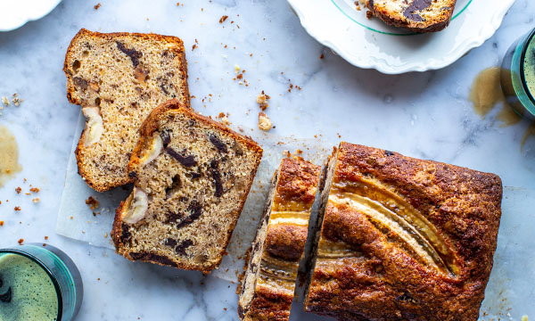Sliced banana bread with swirls of chocolate sits on a marble surface. A whole loaf and two round glasses with green drinks are nearby, evoking a cozy, homemade feel.