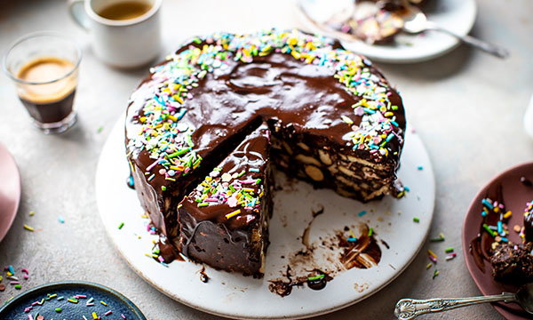 A cake topped with glossy chocolate icing and colourful sprinkles, with a slice cut out, is on a plate. Nearby, cups of coffee and desserts are visible.