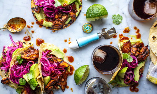 Three tacos with grilled chicken, avocado, and red cabbage coleslaw on a marble surface. Lime wedges and drinks with ice are nearby.