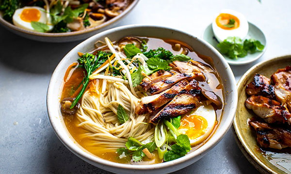 A bowl of ramen with grilled chicken slices, noodles, soft-boiled egg, greens, and herbs in broth. Side dish with egg and herbs is partially visible.