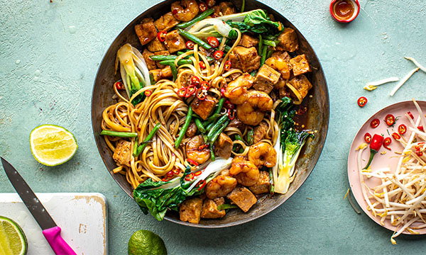 A vibrant bowl of stir-fried noodles with shrimp, tofu, pak choi, and chilli peppers on a textured green surface; lime, knife, and bean sprouts nearby.
