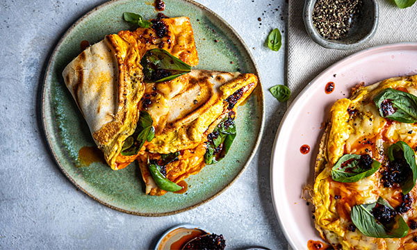 Folded flatbreads omelette with spinach and sun-dried tomatoes on a green plate, alongside a pink plate with more flatbreads. A small dish of black pepper is nearby.