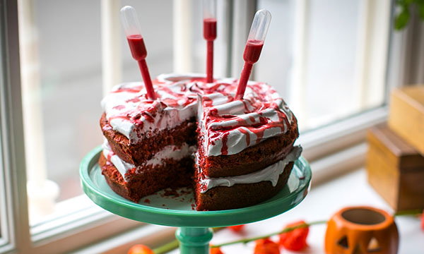 Red velvet Halloween cake with white icing and red syrup drizzle, decorated with pipettes filled with red liquid and displayed on a cake stand