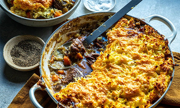Golden shepherd's pie with crispy mashed potato topping in a round dish; filling shows tender beef and vegetables. A bowl of seasoned pie sits nearby.