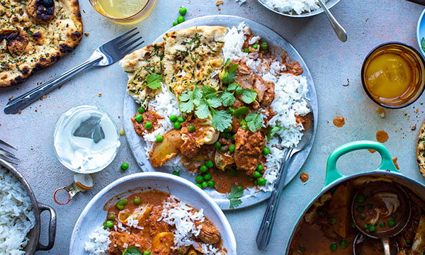 A vibrant Indian meal with curry, rice, naan, and peas on a table. Garnished with coriander, surrounded by drinks and yoghurt, evokes a festive mood.