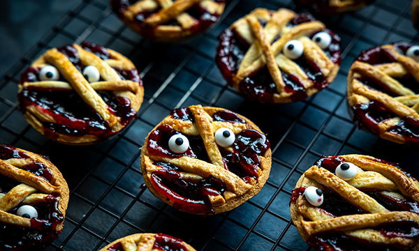 Mini pies with a lattice crust resembling mummy faces, complete with candy eyes, sit on a cooling rack. The pies convey a playful Halloween theme.