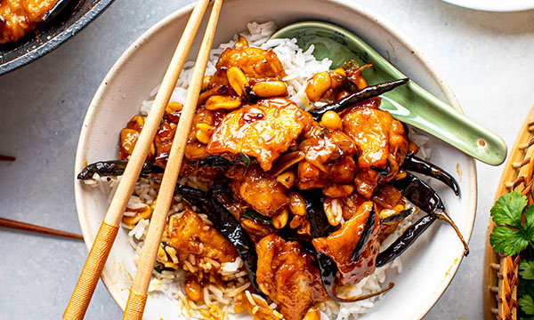 Bowl of Kung Pao chicken served over white rice, topped with roasted peanuts and dried chillies, with chopsticks resting across the bowl