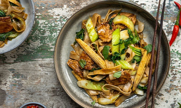 A bowl of stir-fried flat noodles with baby corn, pak choi, and tofu, garnished with sauce. The setting is rustic with chopsticks and chilli.