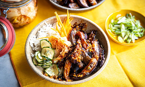 A bowl on a yellow cloth holds rice, cucumber salad, kimchi, and glazed eggplant, creating a vibrant, appetizing display. A small dish of green onions and a jar of kimchi are in the background.