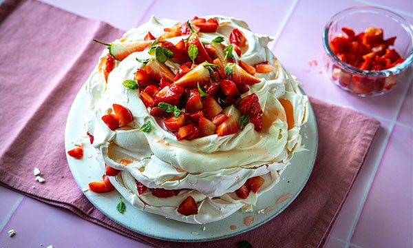 A pavlova topped with fresh strawberries and mint leaves sits on a white plate. The dessert is on a pink napkin, with a bowl of diced strawberries nearby.
