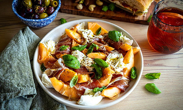 A vibrant salad with slices of cantaloupe, prosciutto, mozzarella, and fresh basil. A glass of iced tea and olives add a rustic, Mediterranean feel.