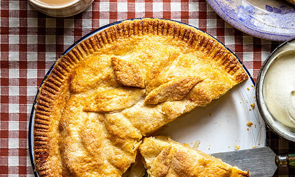 Golden apple tart with a slice removed, on a checked tablecloth. Nearby are a cream bowl and a purple-rimmed plate, creating a cosy, inviting feel.