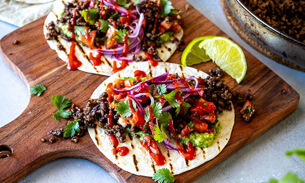 Two tacos on a wooden board, topped with beef, sliced red onions, cilantro, and sauce. A lime wedge is on the side, conveying a fresh, vibrant meal.