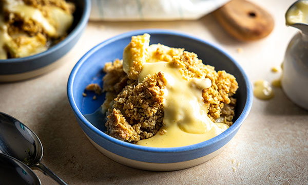 A blue bowl with a serving of apple crumble topped with creamy custard sits on a light surface. Nearby, spoons and another dish enhance the cosy, warm vibe.
