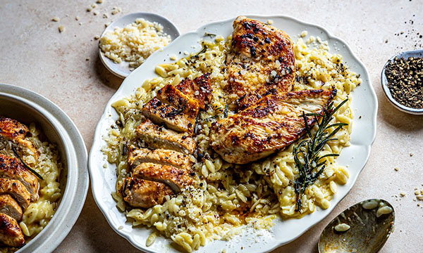 Grilled chicken breast on orzo pasta, garnished with rosemary and parmesan, arranged on a white plate with a rustic, appetising look.